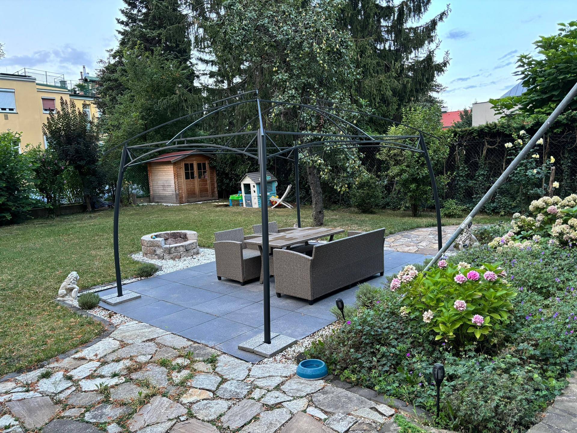 Großer Garten mit gepflasterter Terrasse, Feuerstelle und einem Gartenhaus im Hintergrund.