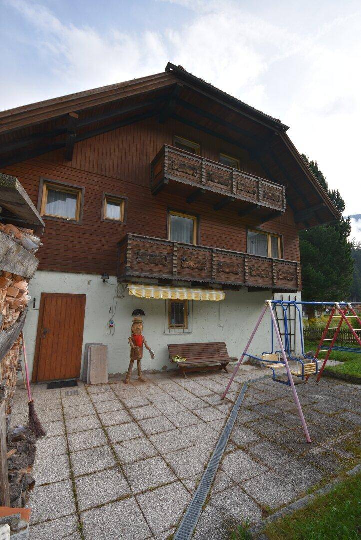 Rückseite des Hauses mit gepflastertem Bereich, Spielplatz und Holzlager.