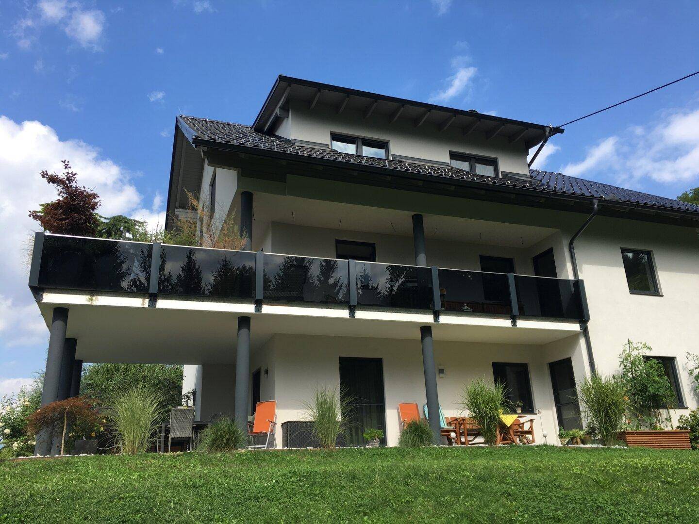 Moderne Hausfassade mit großem Balkon und Glasgeländer unter blauem Himmel.