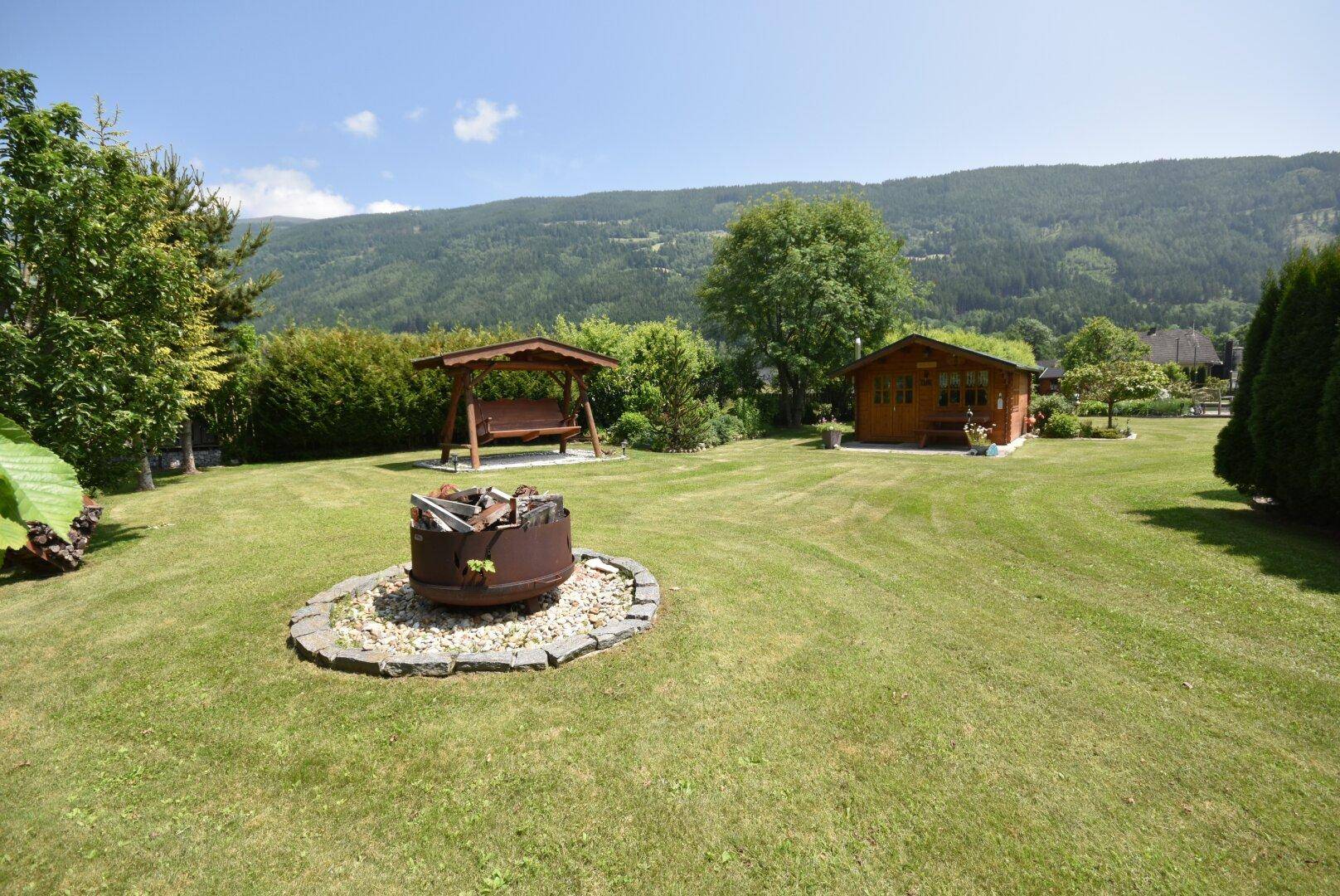 Großer Rasengarten mit Feuerstelle, Holzschaukel und Gartenhaus vor Bergkulisse.