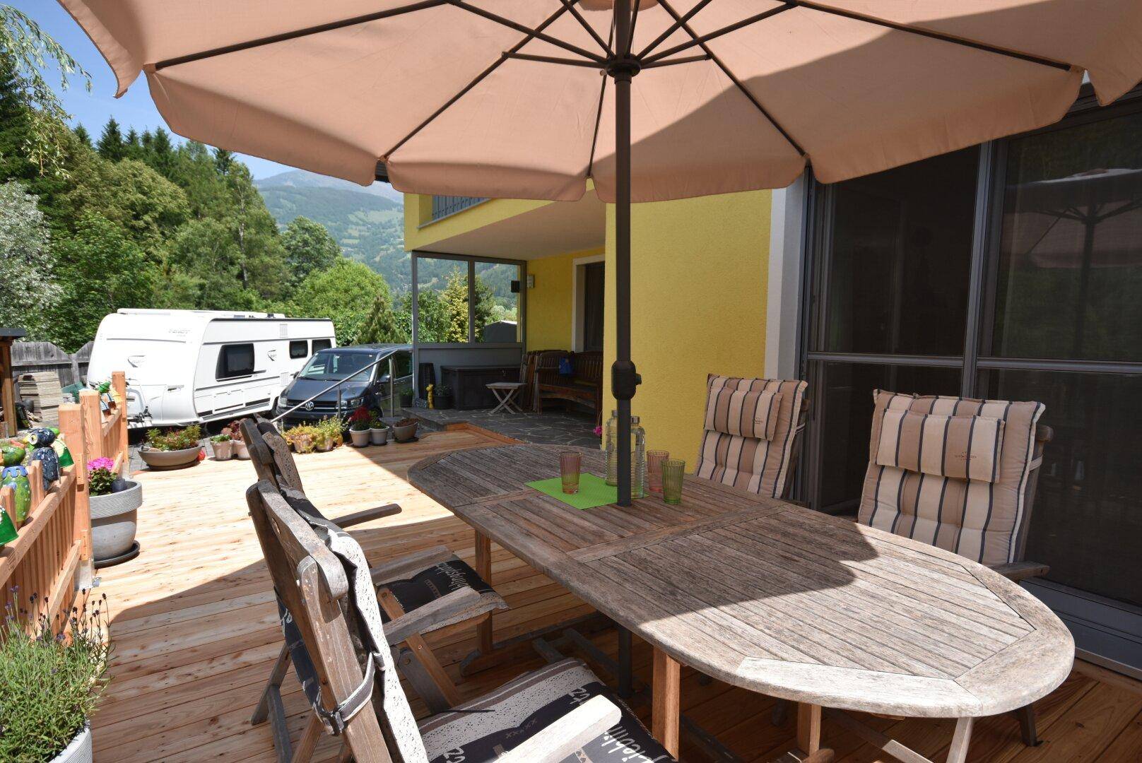 Möblierte Holzterrasse mit großem Sonnenschirm und Esstisch, im Hintergrund ein Wohnwagen und Berge.