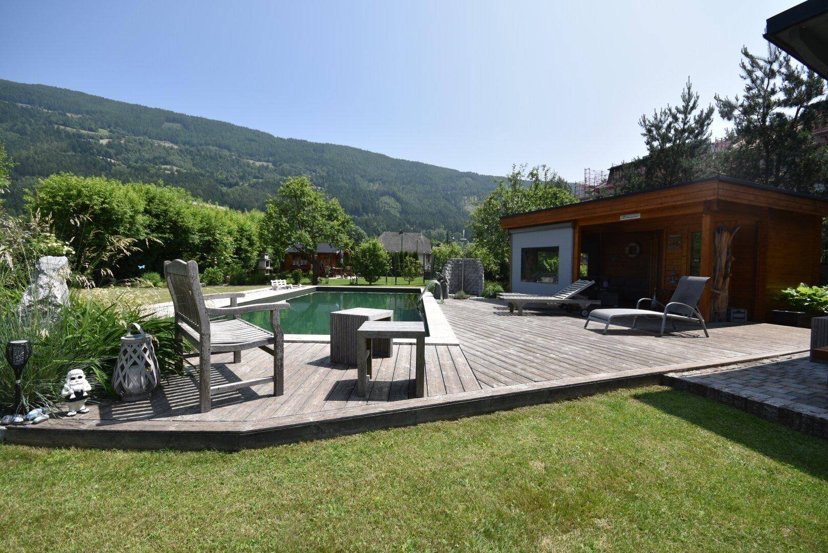 Großer Gartenbereich mit Pool, Holzterrasse und Liegestühlen, umgeben von grünen Bergen.