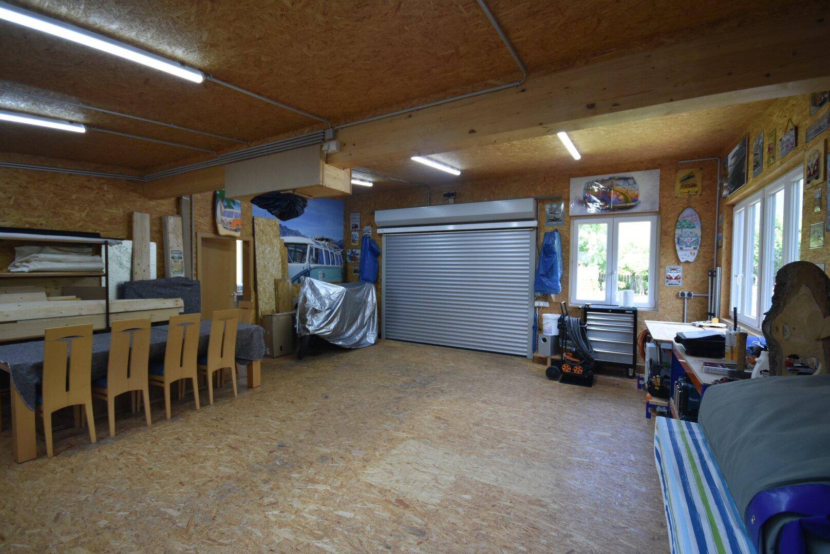 Geräumige Garage oder Werkstatt mit Holzwänden, Rolltor und einem großen Tisch.