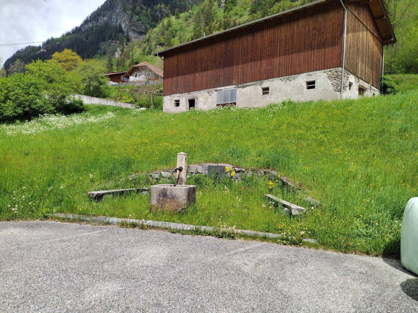 Rustikales Gebäude mit Holzwänden und einem alten Steintrog im Vordergrund, umgeben von Natur.