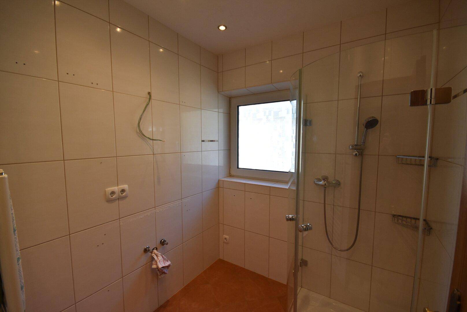 Badezimmer mit Duschbereich, Fliesenwänden und Fenster für Tageslicht.