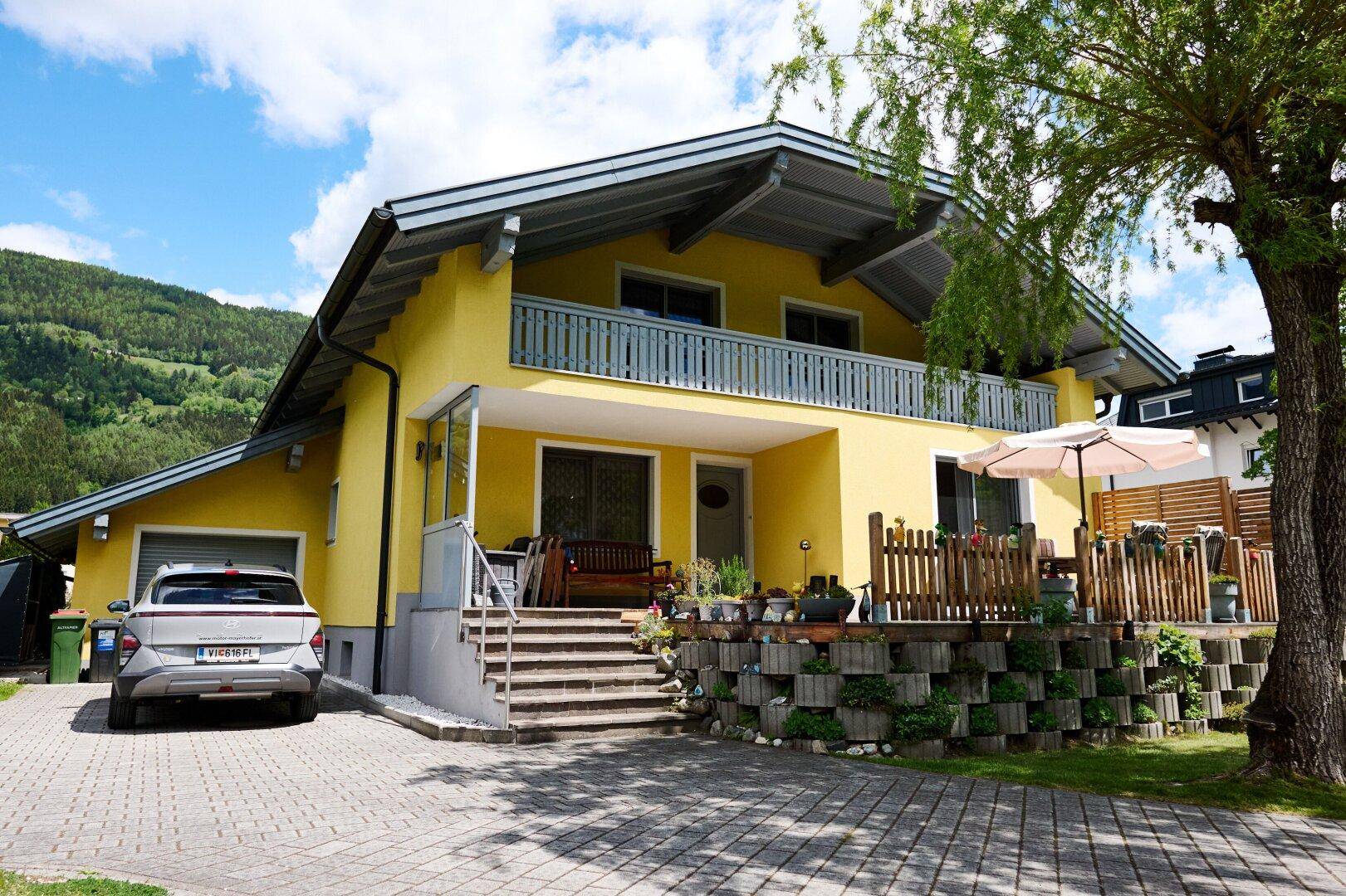 Gelbes Einfamilienhaus mit Balkon, gepflasterter Auffahrt und Bergblick unter blauem Himmel.