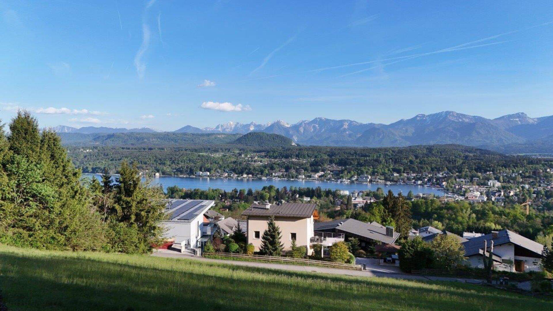 Blick von einer Wiese auf Häuser, einen See und eine beeindruckende Bergkette in der Ferne.