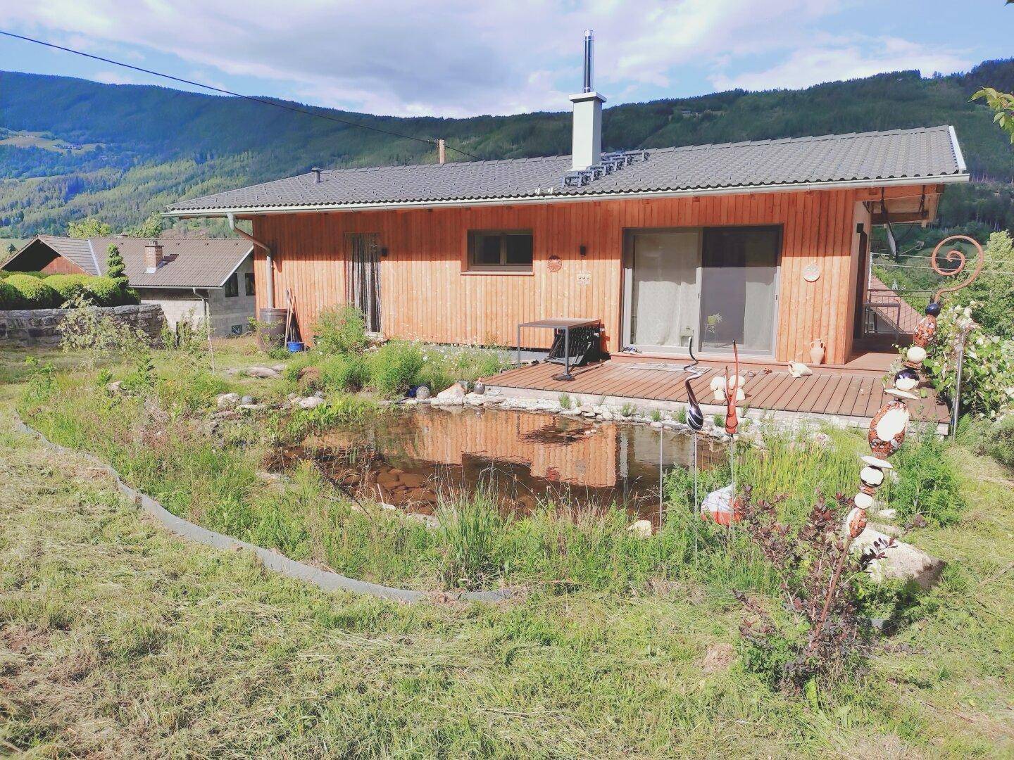 Haus mit Holzfassade, großer Holzterrasse und einem idyllischen Naturteich im Garten.