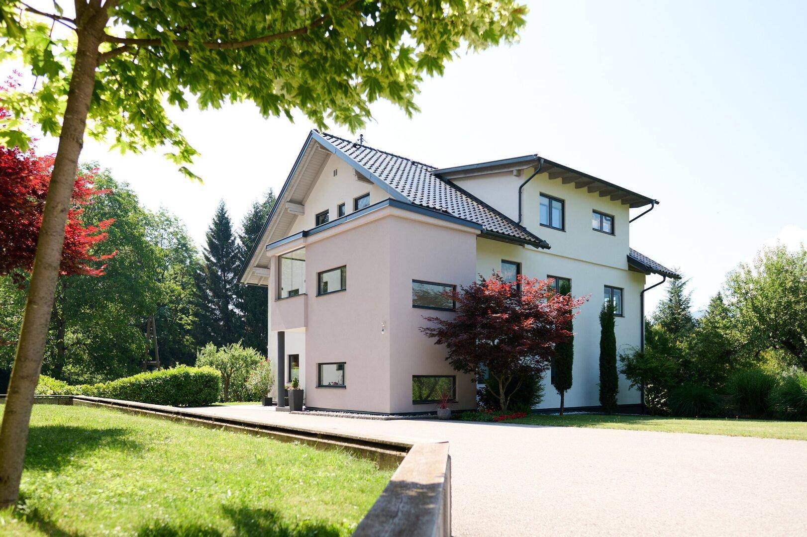 Modernes Wohnhaus mit heller Fassade, dunklem Dach und weitläufiger Auffahrt umgeben von viel Grün.