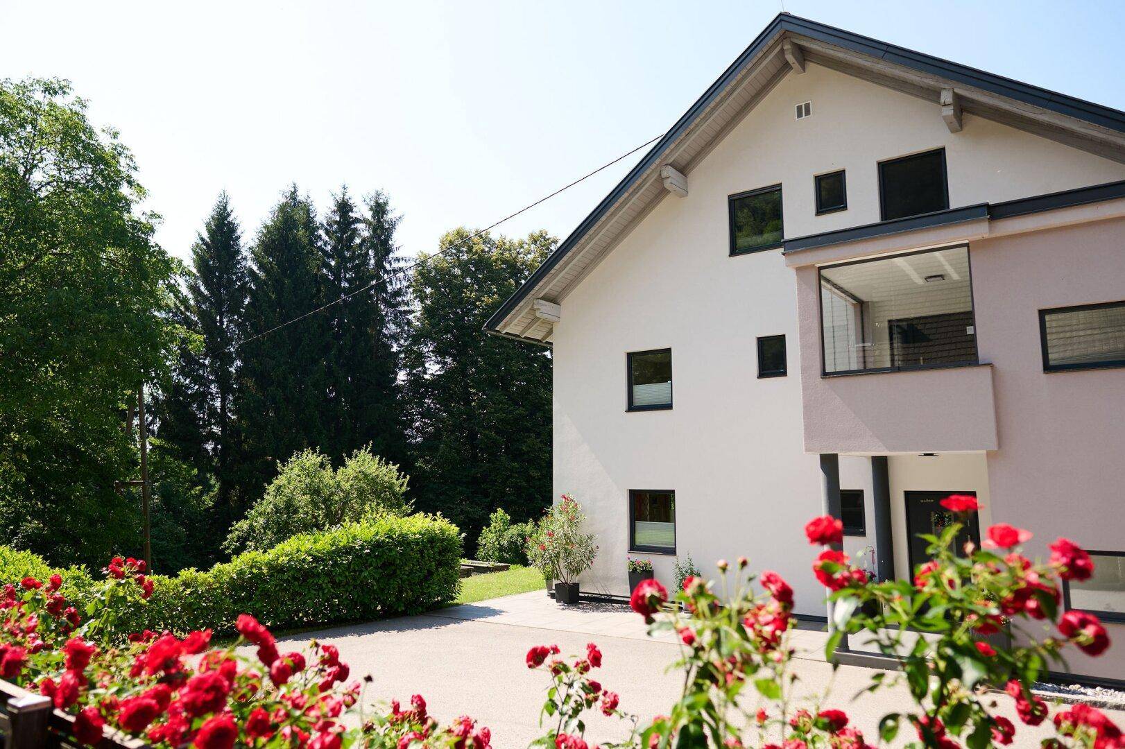 Ansicht des modernen Hauses mit rotem Rosengarten im Vordergrund und hohen Bäumen im Hintergrund.