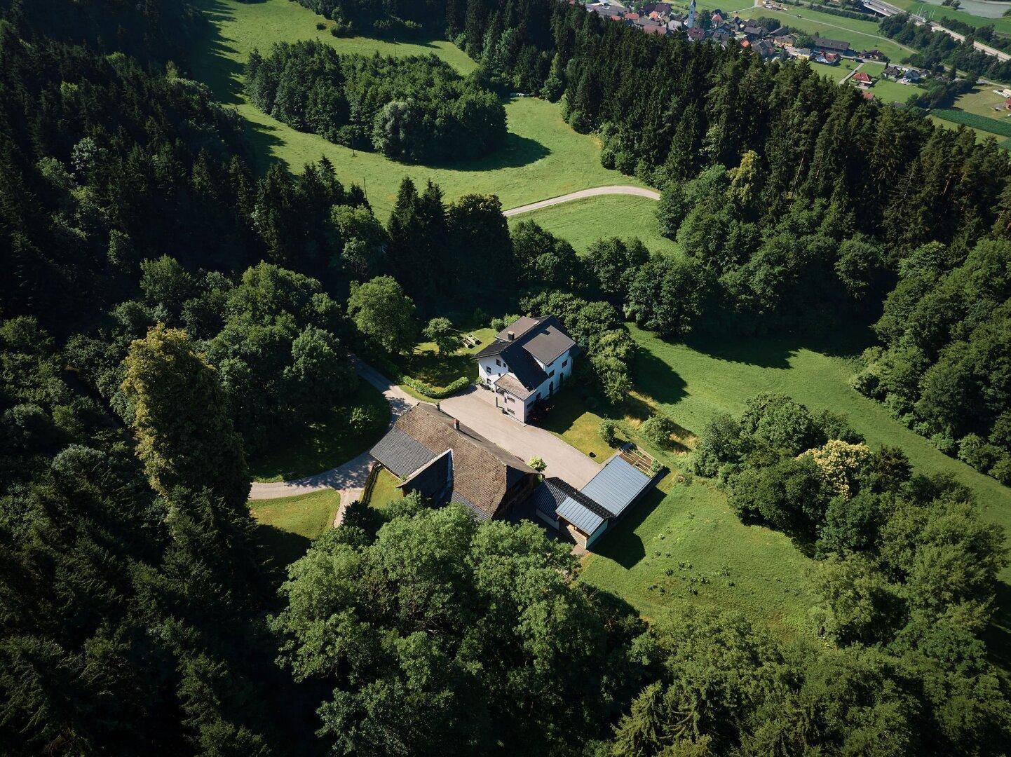 Luftaufnahme eines Anwesens mit zwei Gebäuden, umgeben von dichtem Wald und grünen Wiesen.