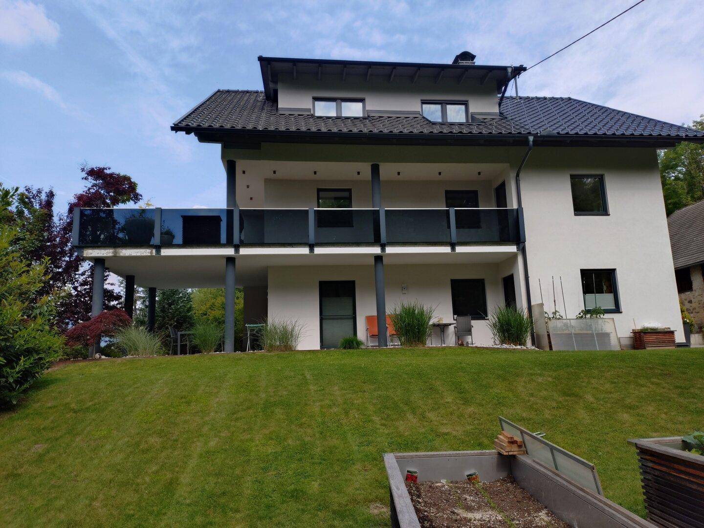 Rückansicht des modernen Hauses mit mehreren Balkonen und einem Hochbeet im sonnigen Gartenbereich.