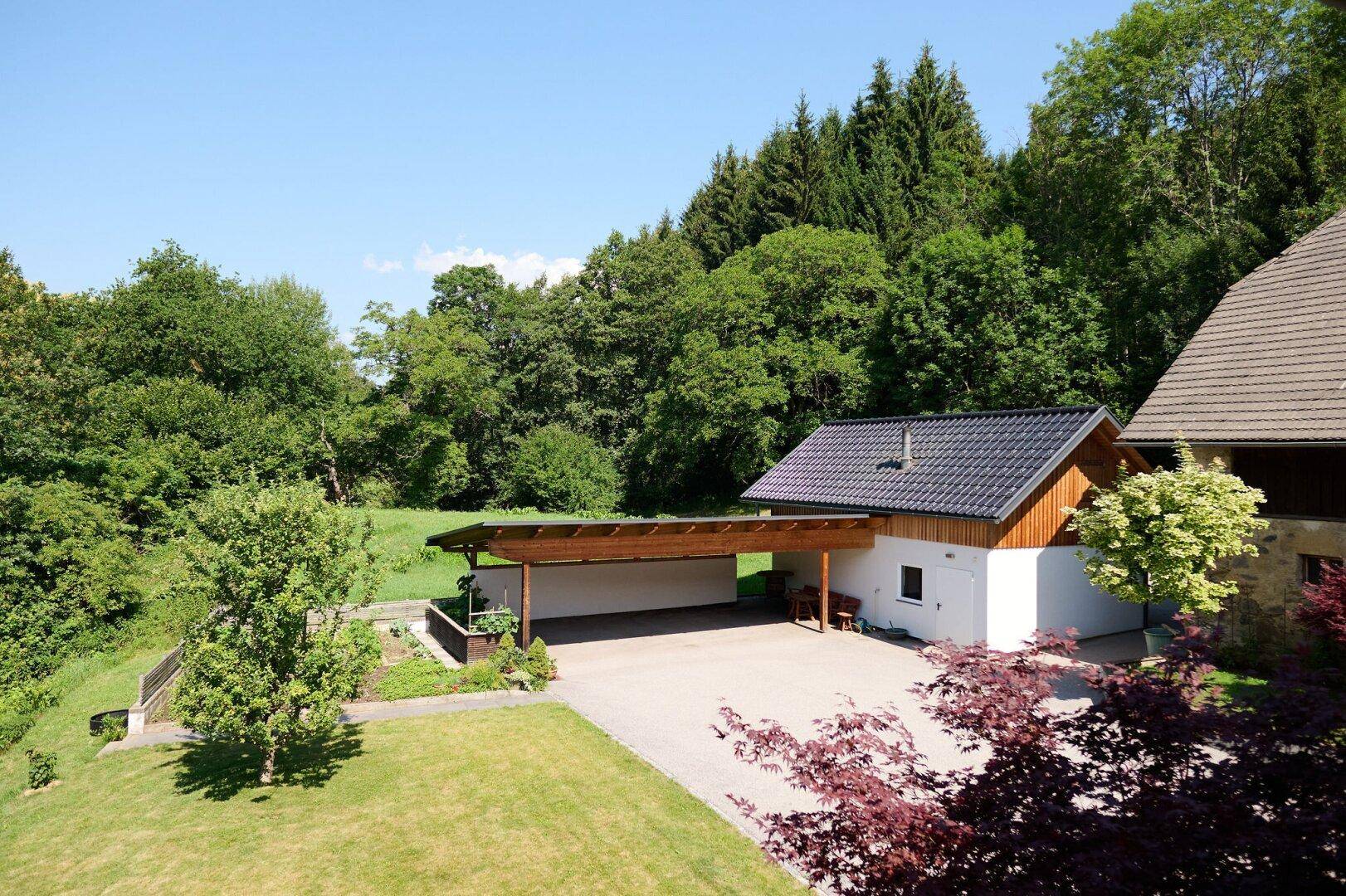 Geräumiger Carport mit Holzdach und gepflastertem Vorplatz, umgeben von üppiger grüner Natur.