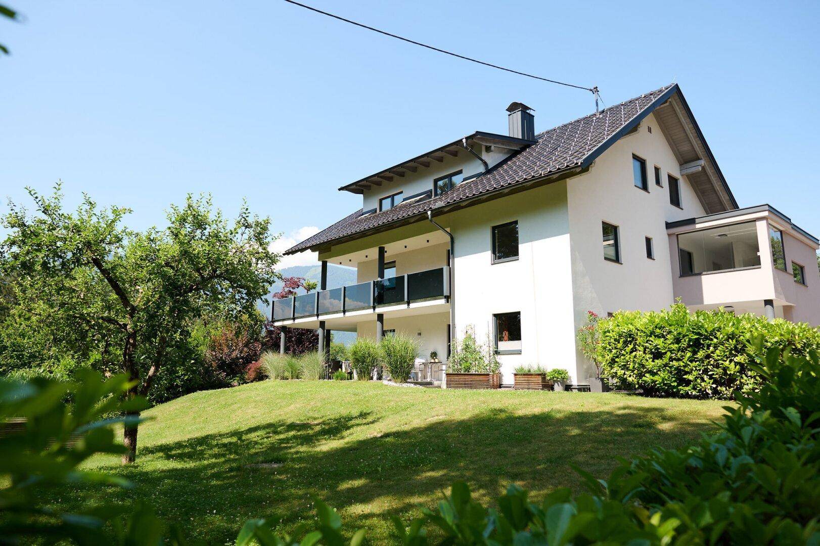 Modernes Wohnhaus mit weißer Fassade, Balkonen mit Glasgeländer und gepflegtem Rasen in sonniger Lage.