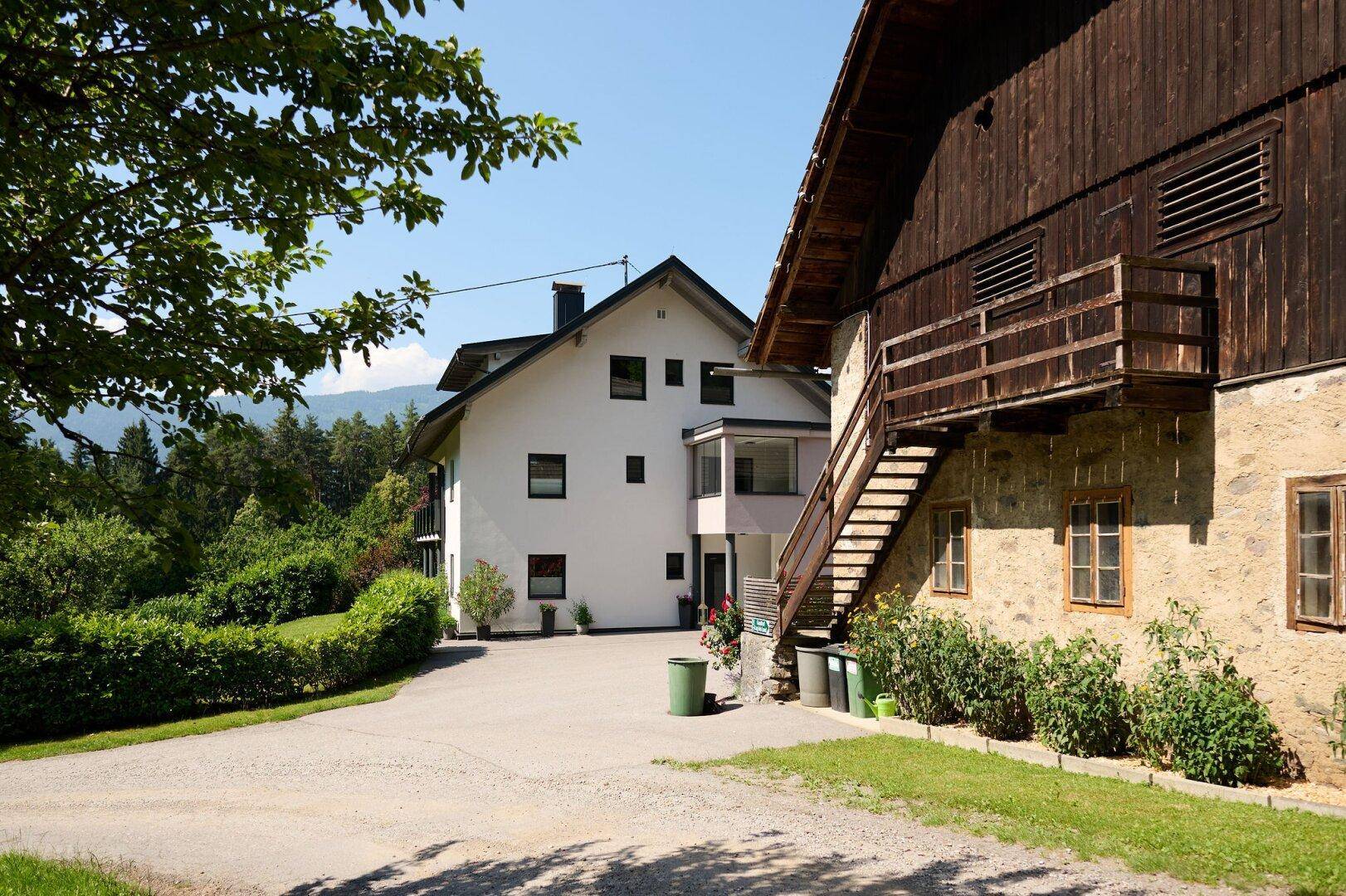 Blick vom Hof auf das moderne weiße Wohnhaus und das angrenzende traditionelle Holz-Stein-Gebäude.