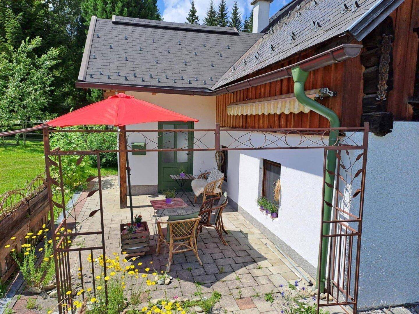 Gemütliche gepflasterte Terrasse mit Gartenmöbeln und rotem Sonnenschirm, umgeben von Blumen.