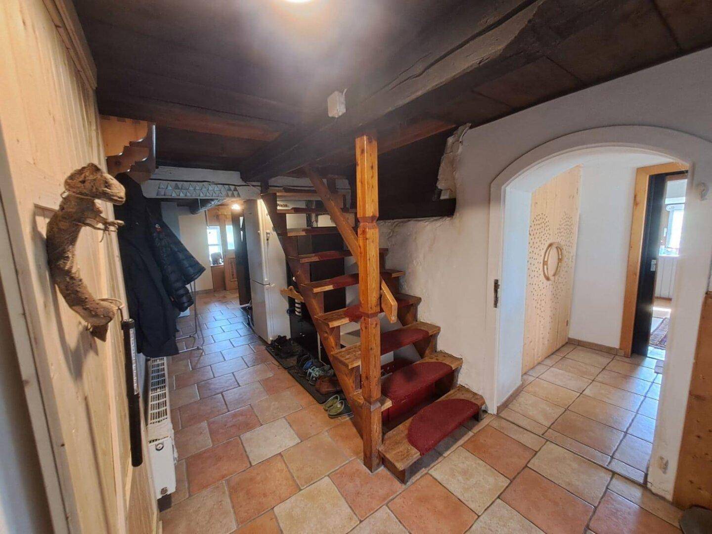Rustikaler Flur mit Holztreppe, sichtbaren Deckenbalken und Terrakotta-Fliesenboden.
