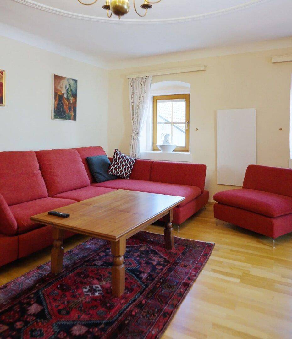Gemütliches Wohnzimmer mit rotem Sofa, Holztisch und Parkettboden unter einer Stuckdecke.