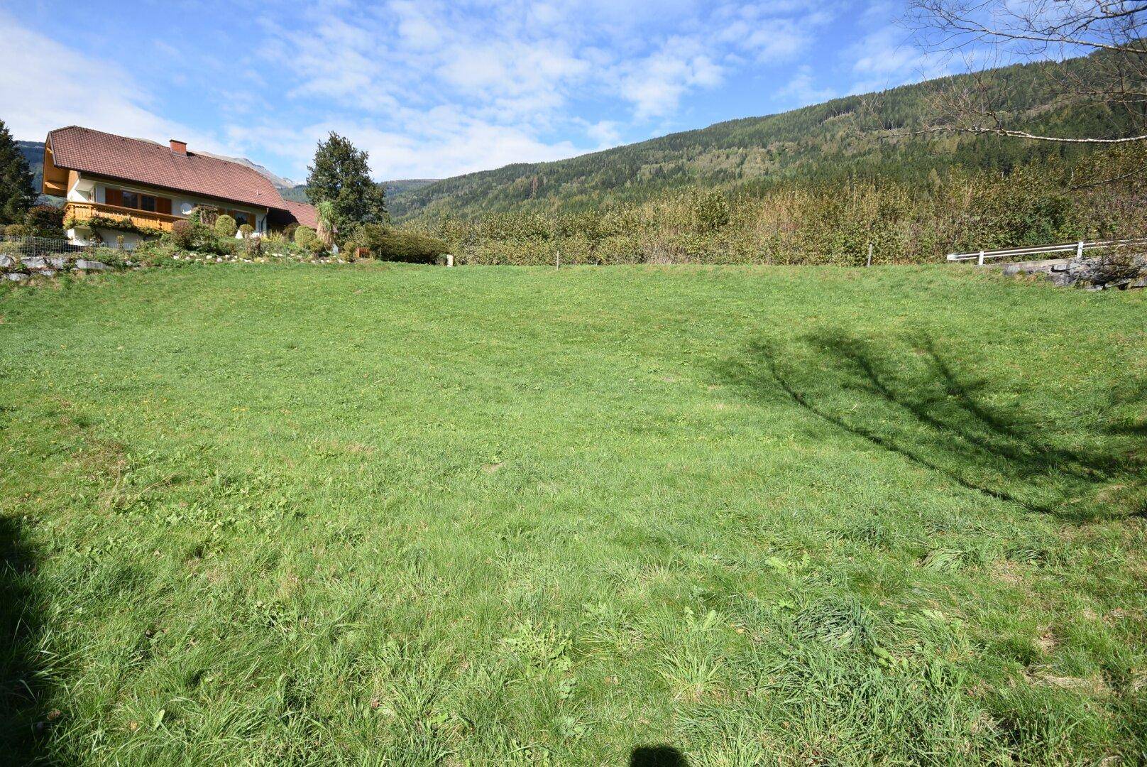 Weitläufige grüne Wiese mit sanftem Hang und einem Haus am Horizont unter blauem Himmel.
