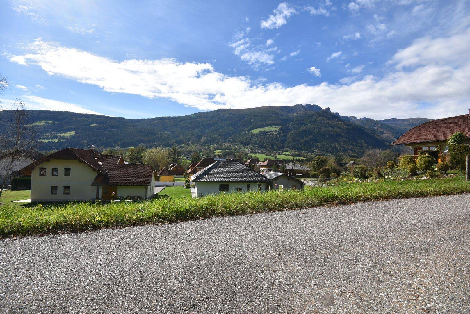 Blick von einem Schotterweg auf ein Wohngebiet mit Häusern und einer malerischen Bergkulisse.