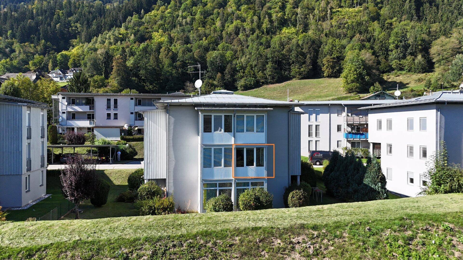 Mehrfamilienhaus in grüner Umgebung mit Balkonen und Blick auf bewaldete Hügel.