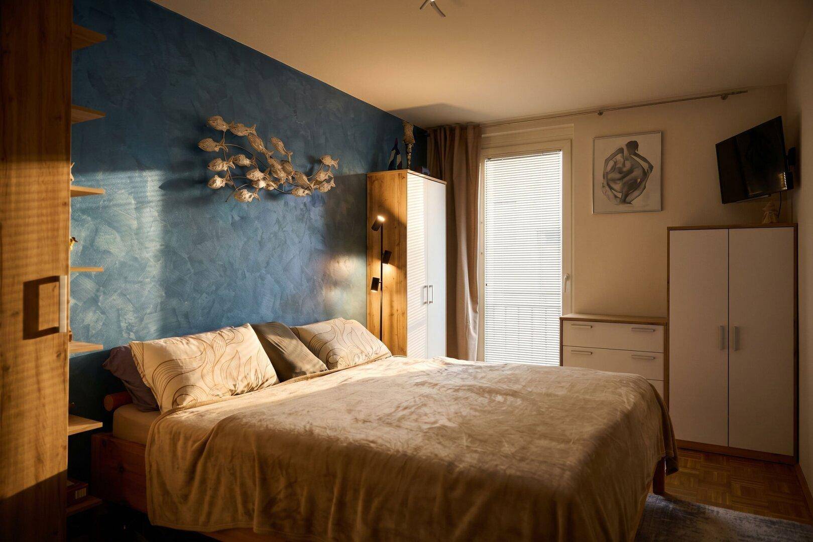 Helles Schlafzimmer mit blauer Wand, großem Bett und Fenster für natürliches Licht.