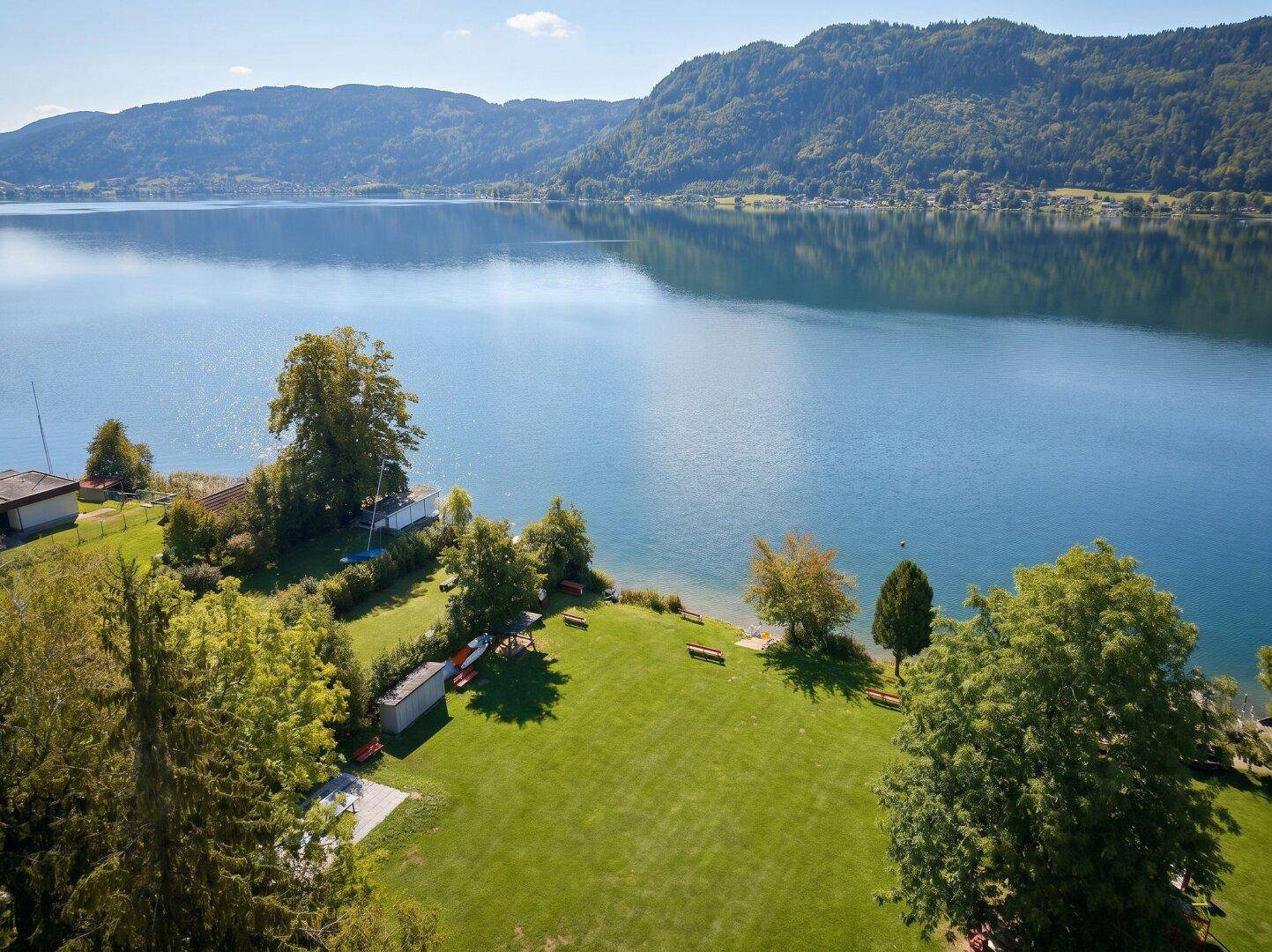 Weitläufige grüne Liegewiese am Seeufer mit schattenspendenden Bäumen und Bergblick.
