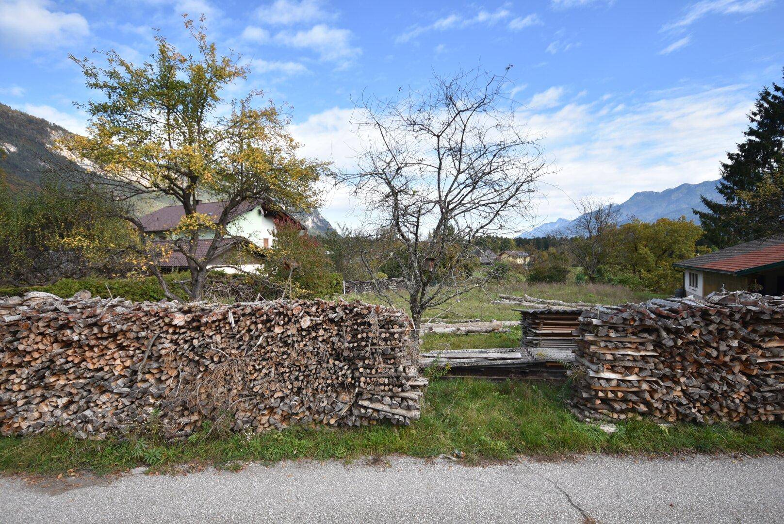 Grundstückszugang von der Straße, flankiert von großen Holzstapeln mit Bergblick im Hintergrund.