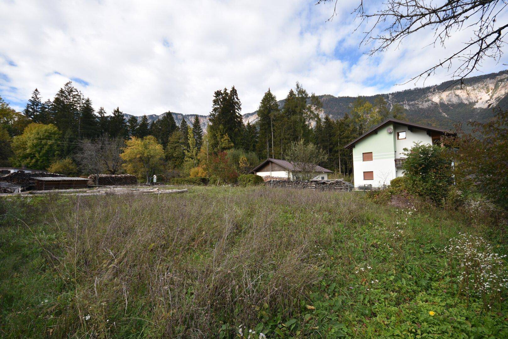 Freies Grundstück mit Blick auf ein Wohnhaus, umgeben von Bäumen und einer beeindruckenden Bergkette.