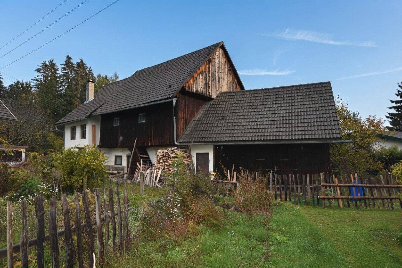 Altes Holzgebäude mit Anbau und dunklem Ziegeldach, umgeben von einem rustikalen Holzzaun.