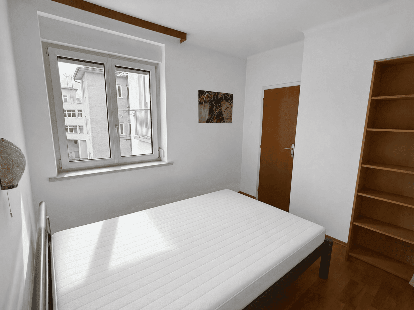 Schlafzimmer mit Holzboden, einem Bett und einem Bücherregal, hell durch das Fenster.