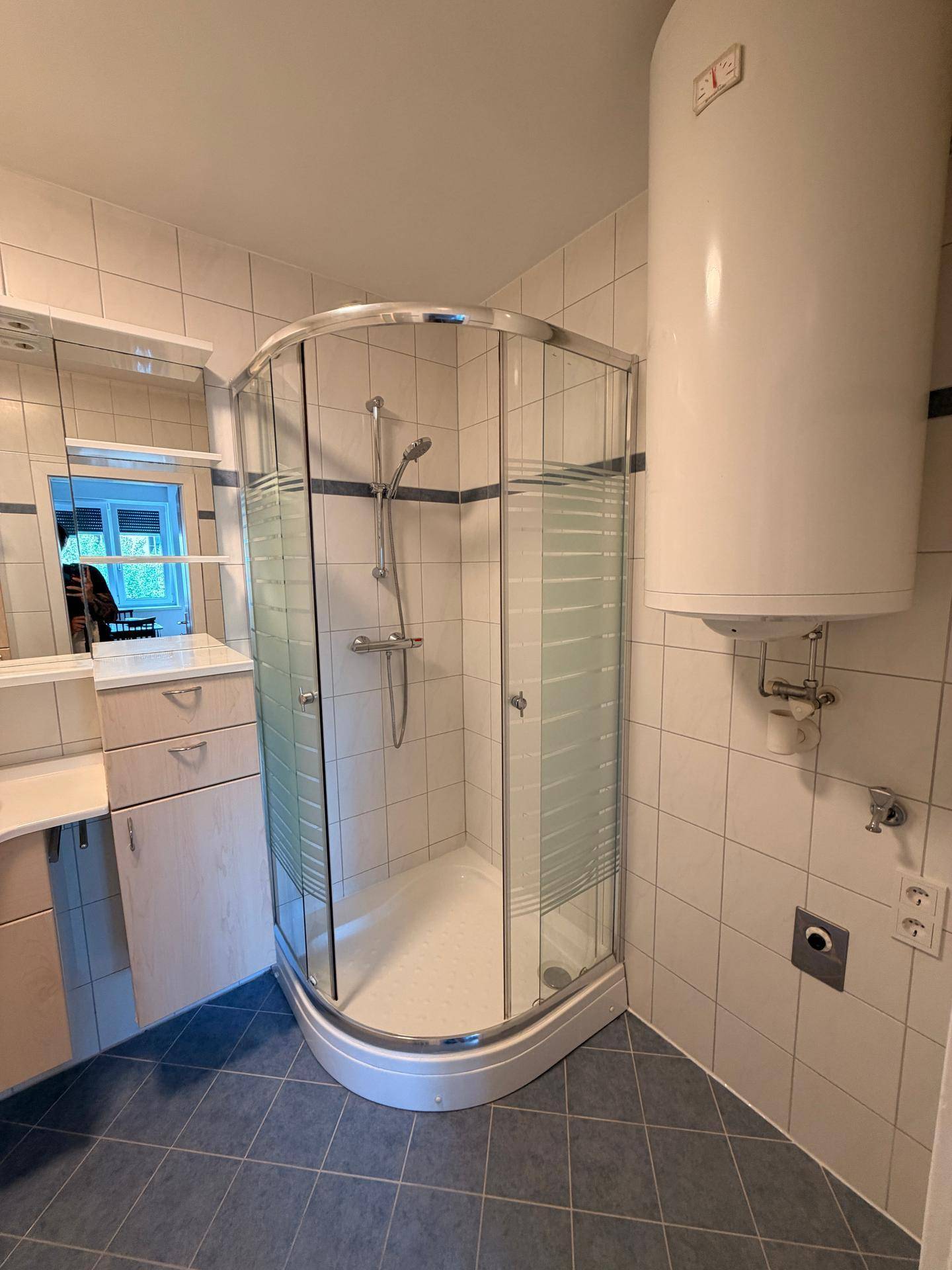 Badezimmer mit Duschkabine und Warmwasserboiler, gefliest in hellen und blauen Tönen.
