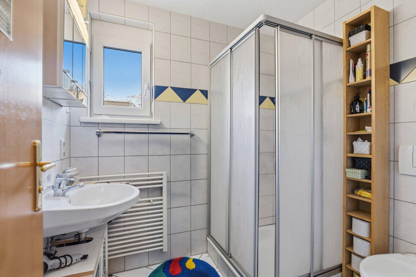 Badezimmer mit Duschkabine, Fenster, Waschbecken und Handtuchheizkörper.