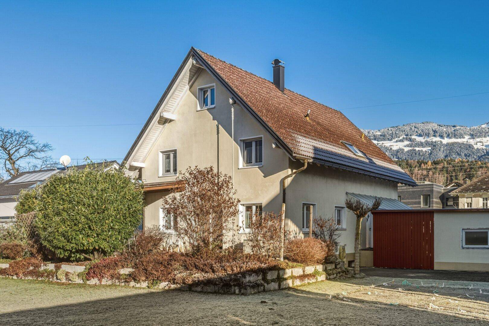 Das Einfamilienhaus mit Satteldach und Garten, umgeben von Bergen unter strahlend blauem Himmel.