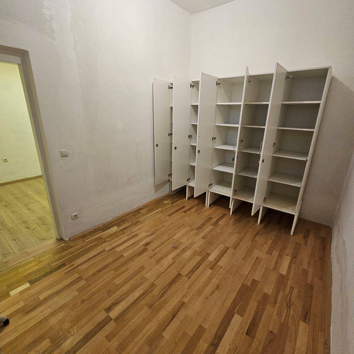Raum mit Holzparkettboden und offenen Regalschränken, die Stauraum bieten, mit Blick in einen Nebenraum.