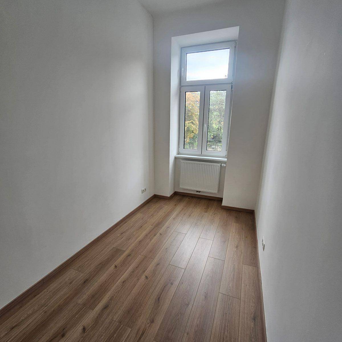 Schmaler Raum mit Laminatboden und einem hohen Fenster, ideal als Schlaf- oder Arbeitszimmer.