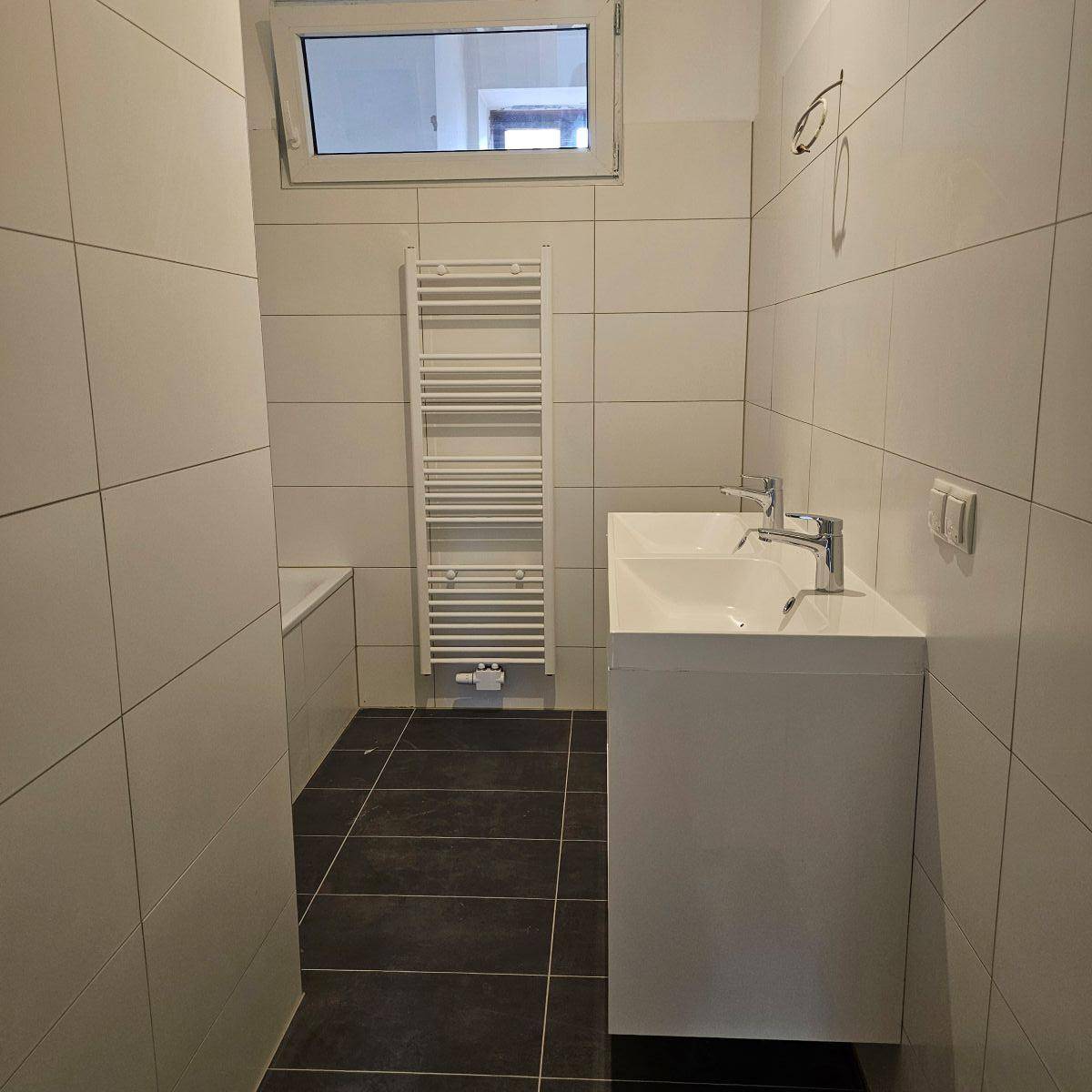 Modernes Badezimmer mit Doppelwaschbecken, weißen Fliesen und dunklem Boden, inklusive Handtuchheizkörper.