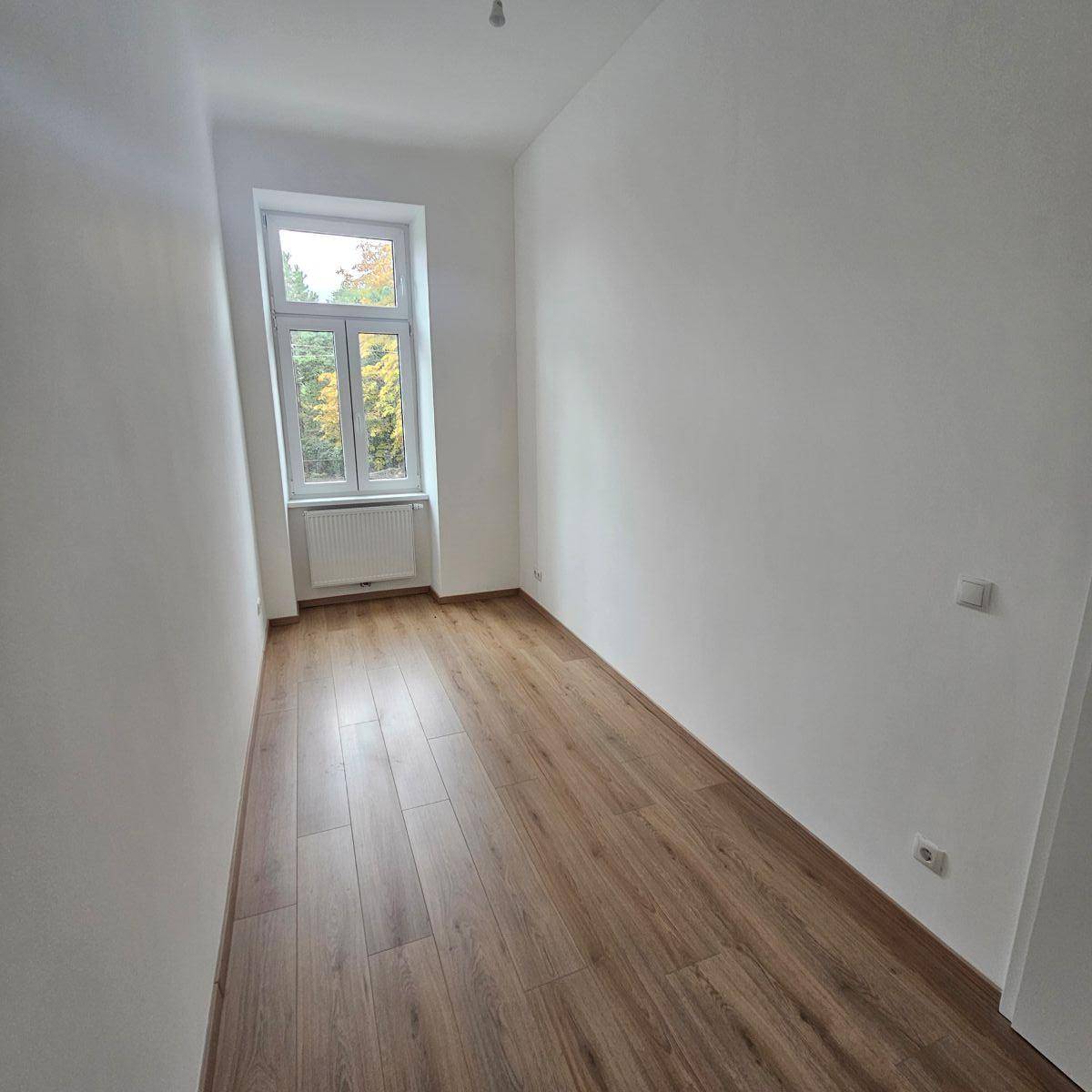 Heller, leerer Raum mit Laminatboden und einem hohen Fenster, das eine grüne Aussicht bietet.