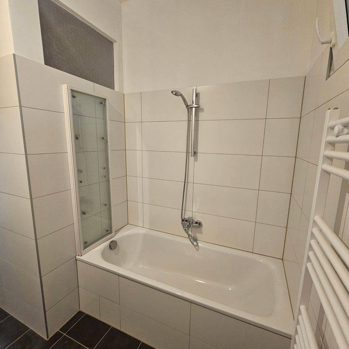 Badezimmer mit Badewanne und Duschabtrennung, weißen Wandfliesen und dunklem Boden.