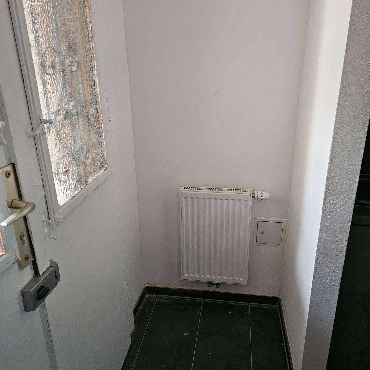 Eingangsbereich mit dunklem Fliesenboden, Heizkörper und einer Tür mit Glaseinsatz.
