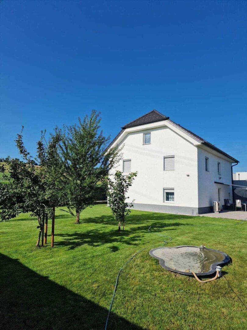 Weißes Einfamilienhaus mit großem, gepflegtem Rasen und jungen Bäumen an einem sonnigen Tag.