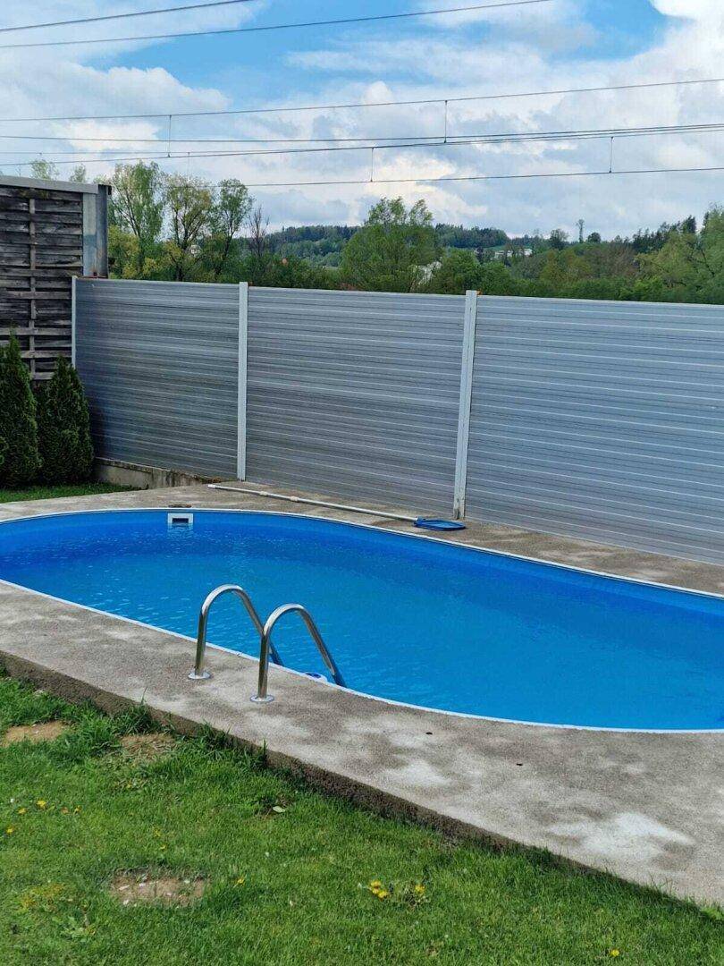 Ovaler Swimmingpool im Garten mit Metallzaun und grüner Umgebung unter blauem Himmel.