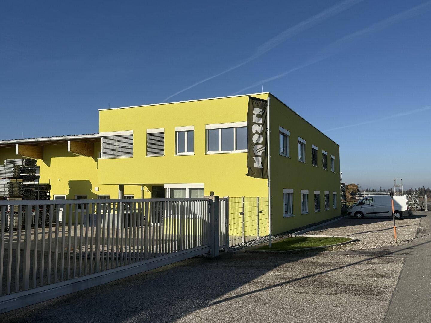 Modernes gelbes Geschäftsgebäude mit großen Fenstern, Zaun und Parkmöglichkeiten im Freien.