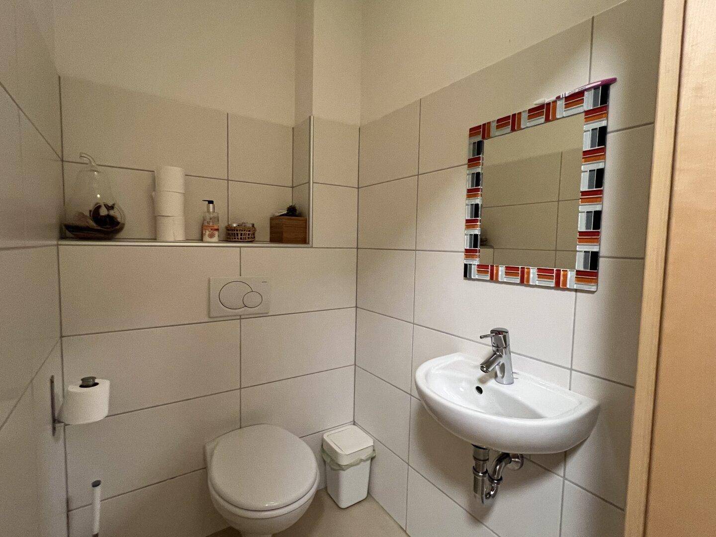 Kleines Gäste-WC mit weißen Fliesen, Wand-WC und farbigem Spiegel.