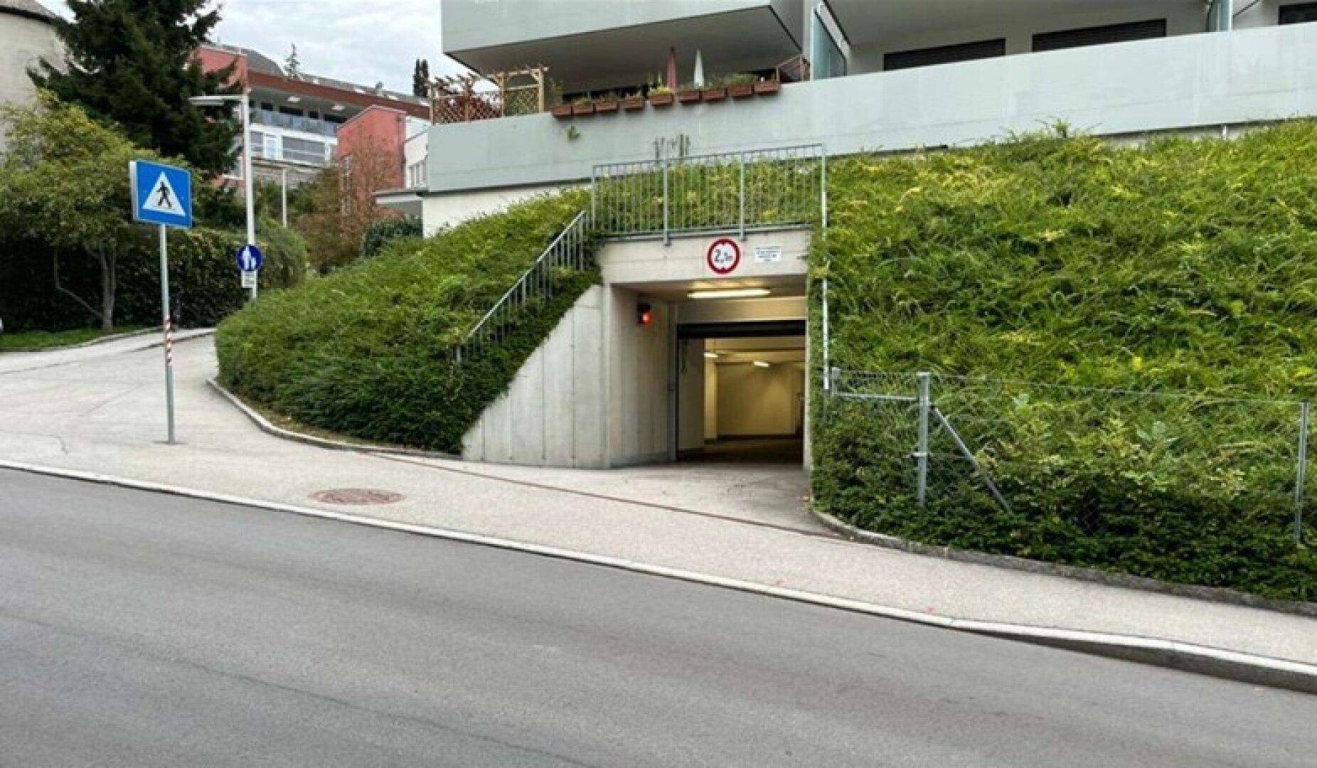 Einfahrt zur Tiefgarage, umgeben von grünen Büschen und einer Wohnanlage im Hintergrund.