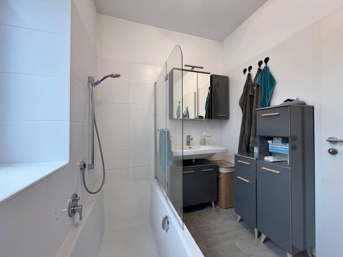 Modernes Badezimmer mit Badewanne, Duschfunktion, Waschbecken und praktischen grauen Schränken.