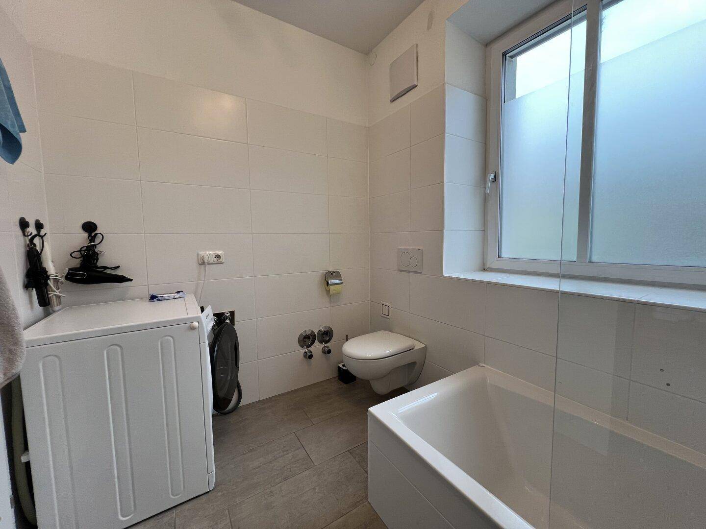 Funktionales Badezimmer mit Badewanne, Toilette, Waschmaschine und einem Fenster für Tageslicht.