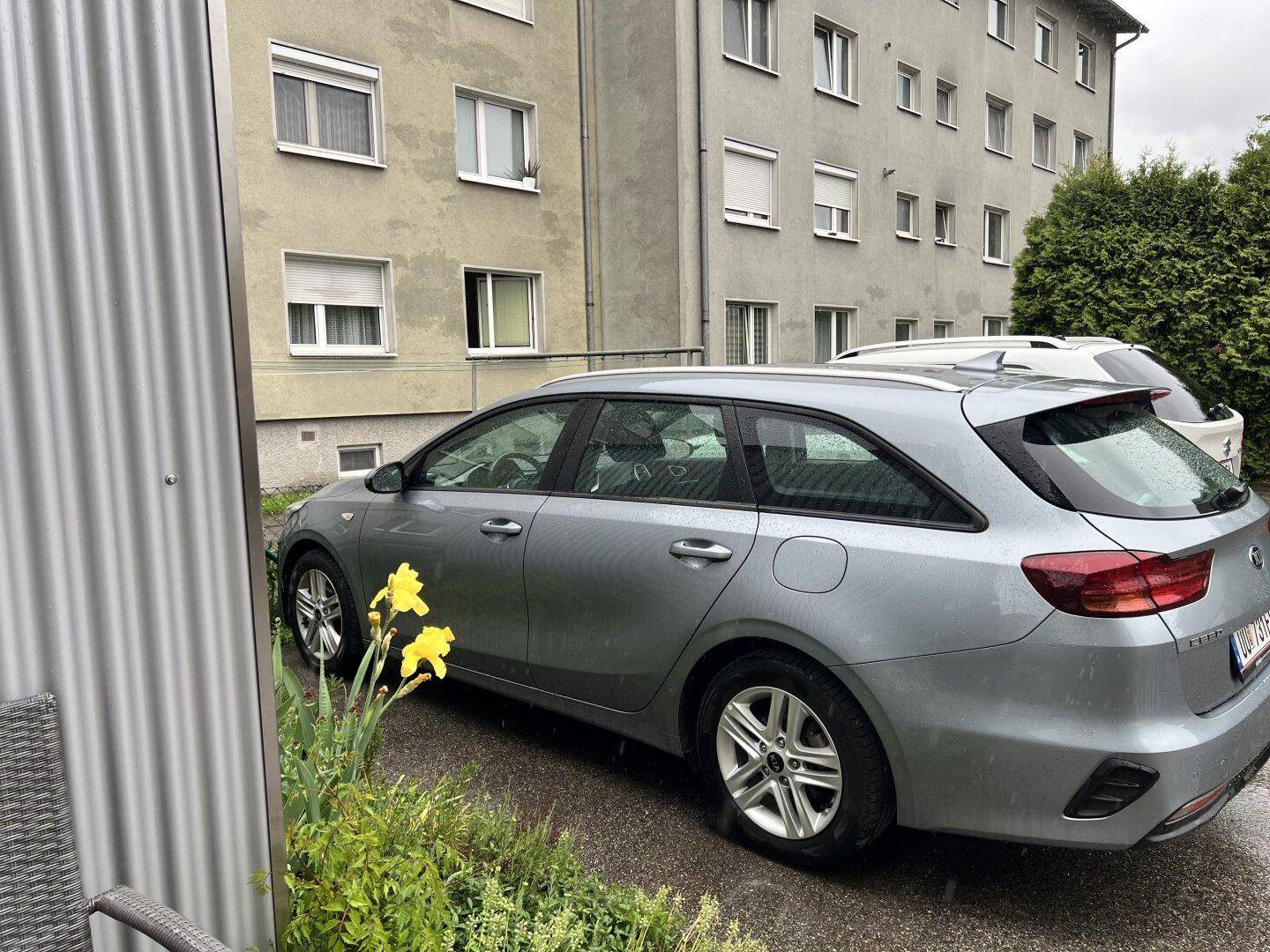 Graues Auto parkt vor einem Mehrfamilienhaus mit mehreren Fenstern und grünen Pflanzen.