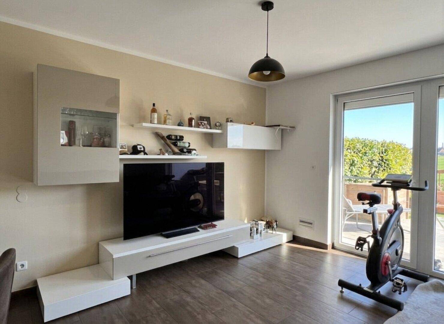 Helles Wohnzimmer mit modernem TV-Möbel, Trainingsrad und Zugang zu einem Außenbereich.