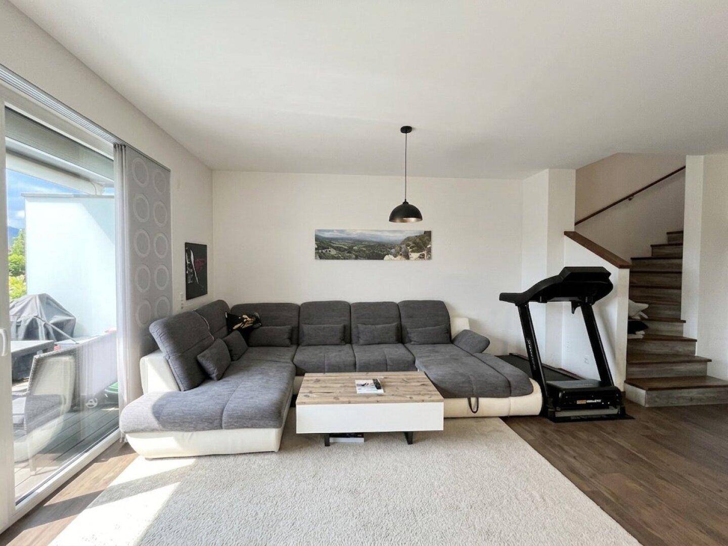 Geräumiges Wohnzimmer mit großer Eckcouch, Laufband und Zugang zu einem Balkon oder einer Terrasse.